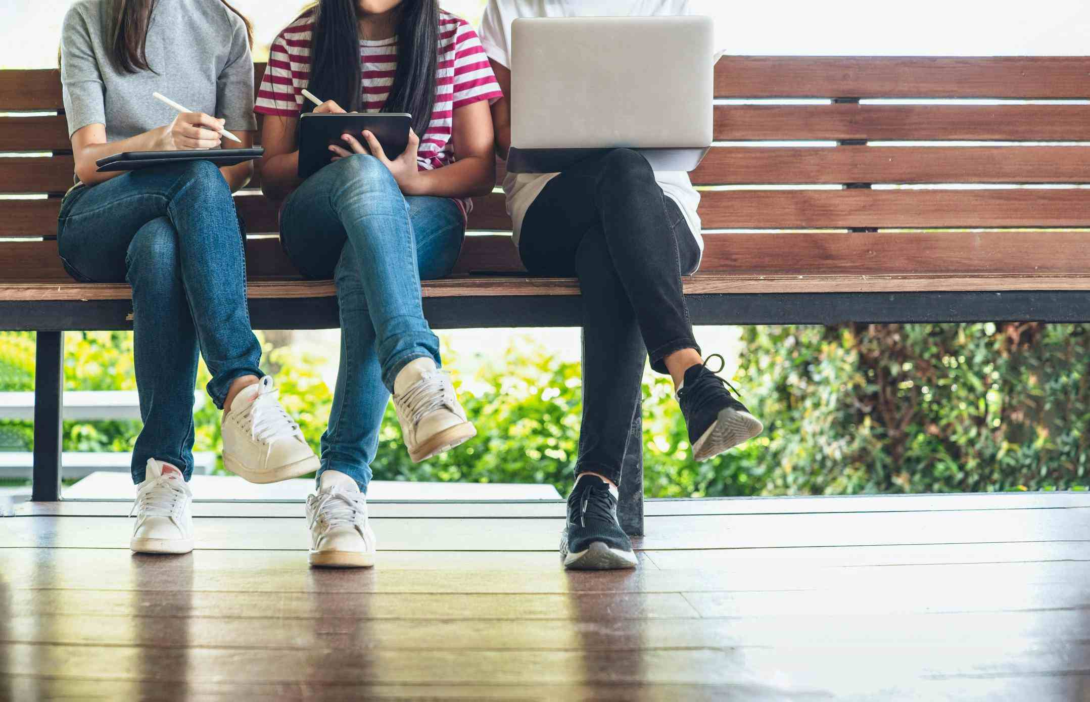 Three female students sitting on a bench together working on their tablets and laptops