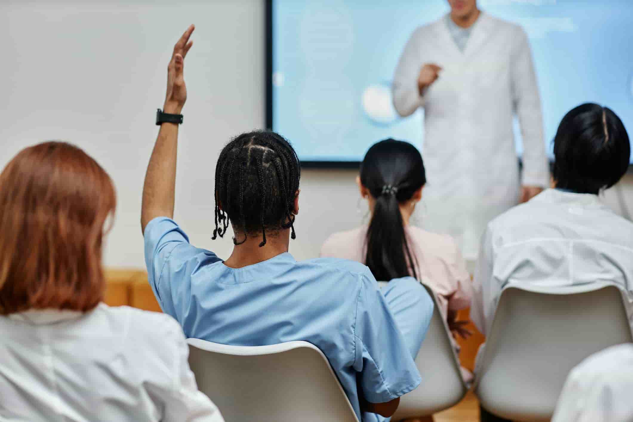 Medical student raising hand in classroom during a lecture with a professor in a white coat presenting in front of a screen. Diverse group of students wearing scrubs attentively listening and engaging in a healthcare education setting.