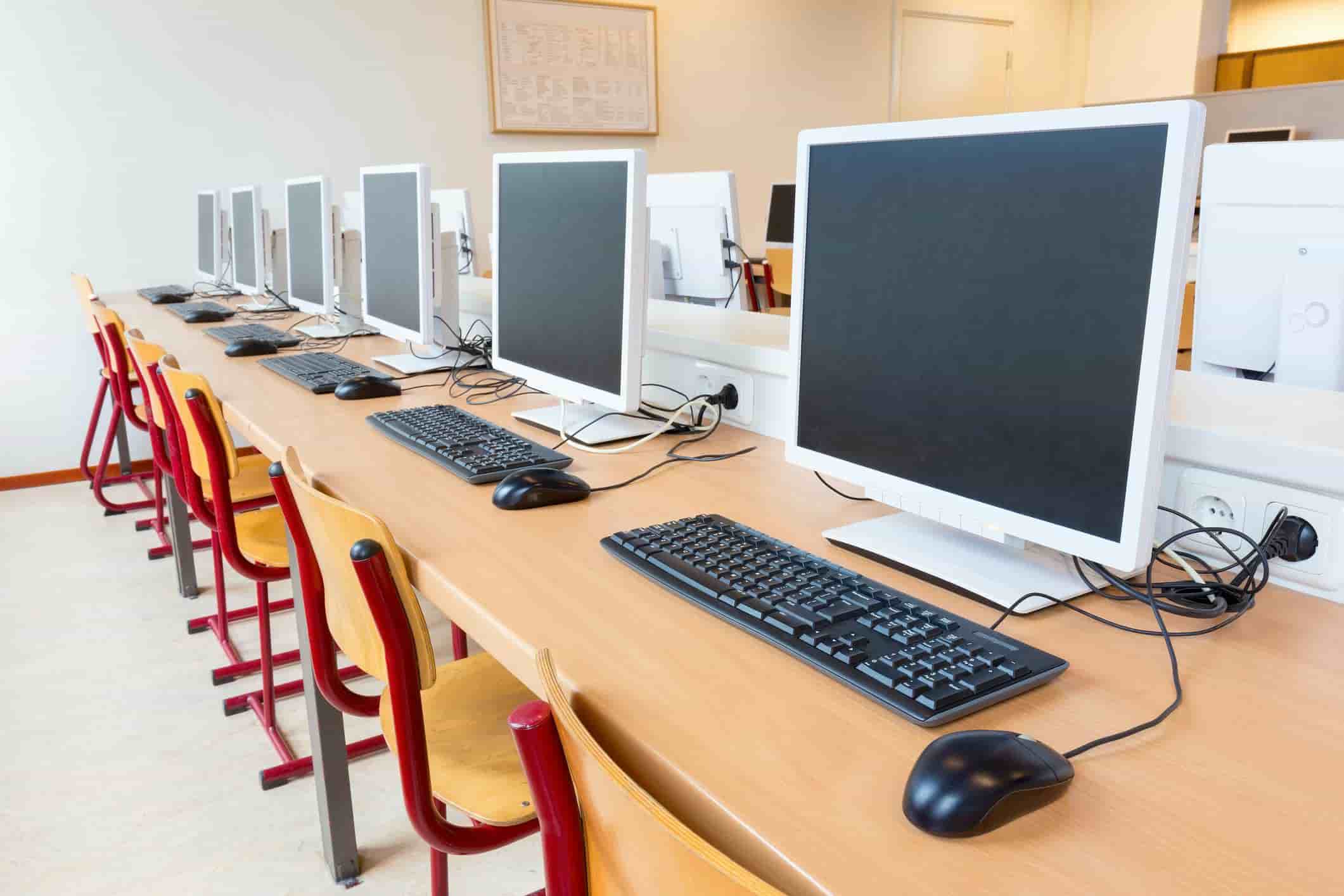 Empty computer lab with a row of desktop computers on wooden desks, black keyboards, and mice, in a brightly lit classroom. Red and wooden chairs are neatly arranged at each station, with power outlets visible on the desk. The room has a clean, modern setup designed for educational or training purposes.