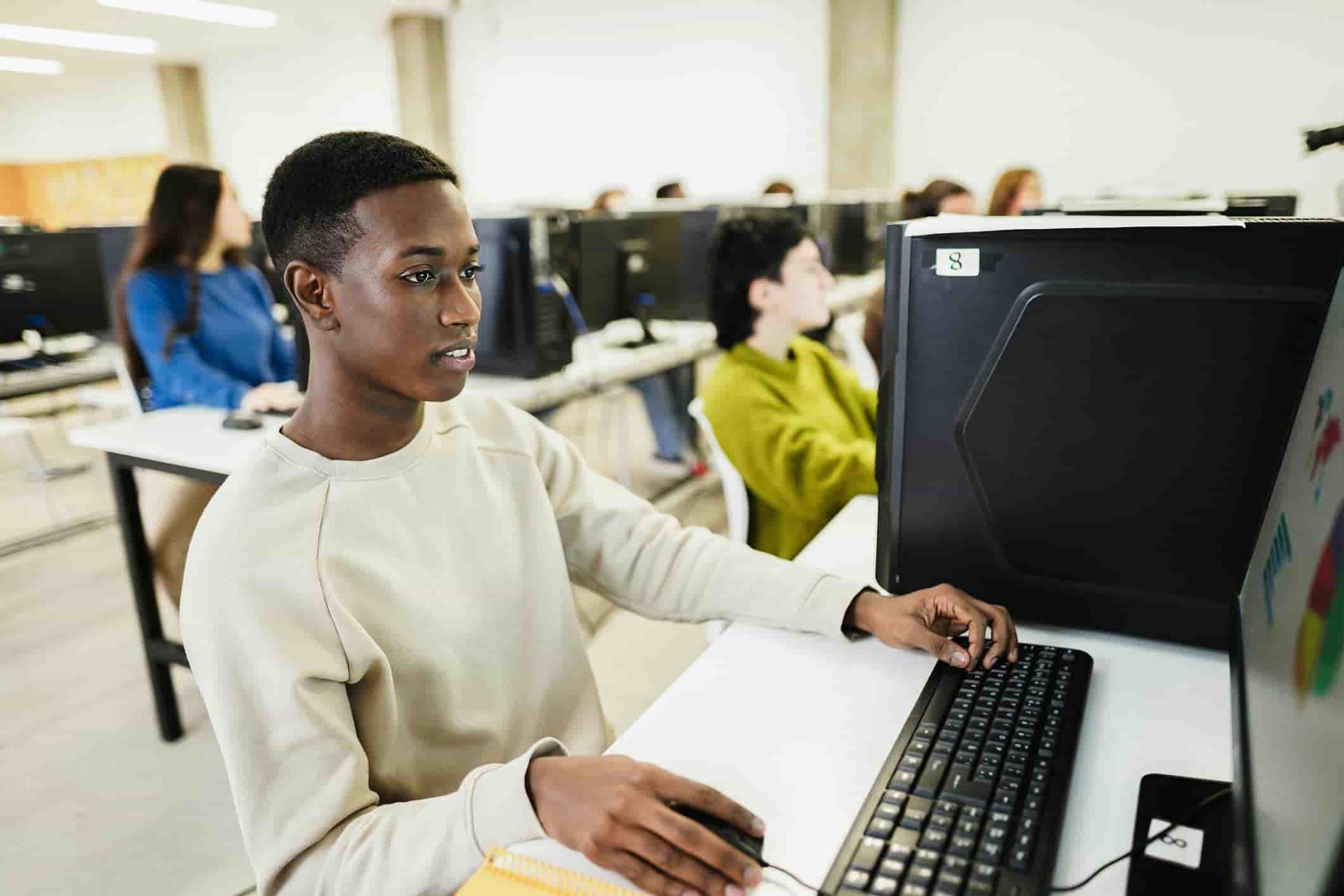 Male high school student focused on a computer screen while working in a computer lab, with other students in the background, during digital ACT. He is using a desktop computer, wearing a light beige sweatshirt, and holding a mouse. The setting appears to be a modern classroom with several students at their desks engaged in digital ACT.