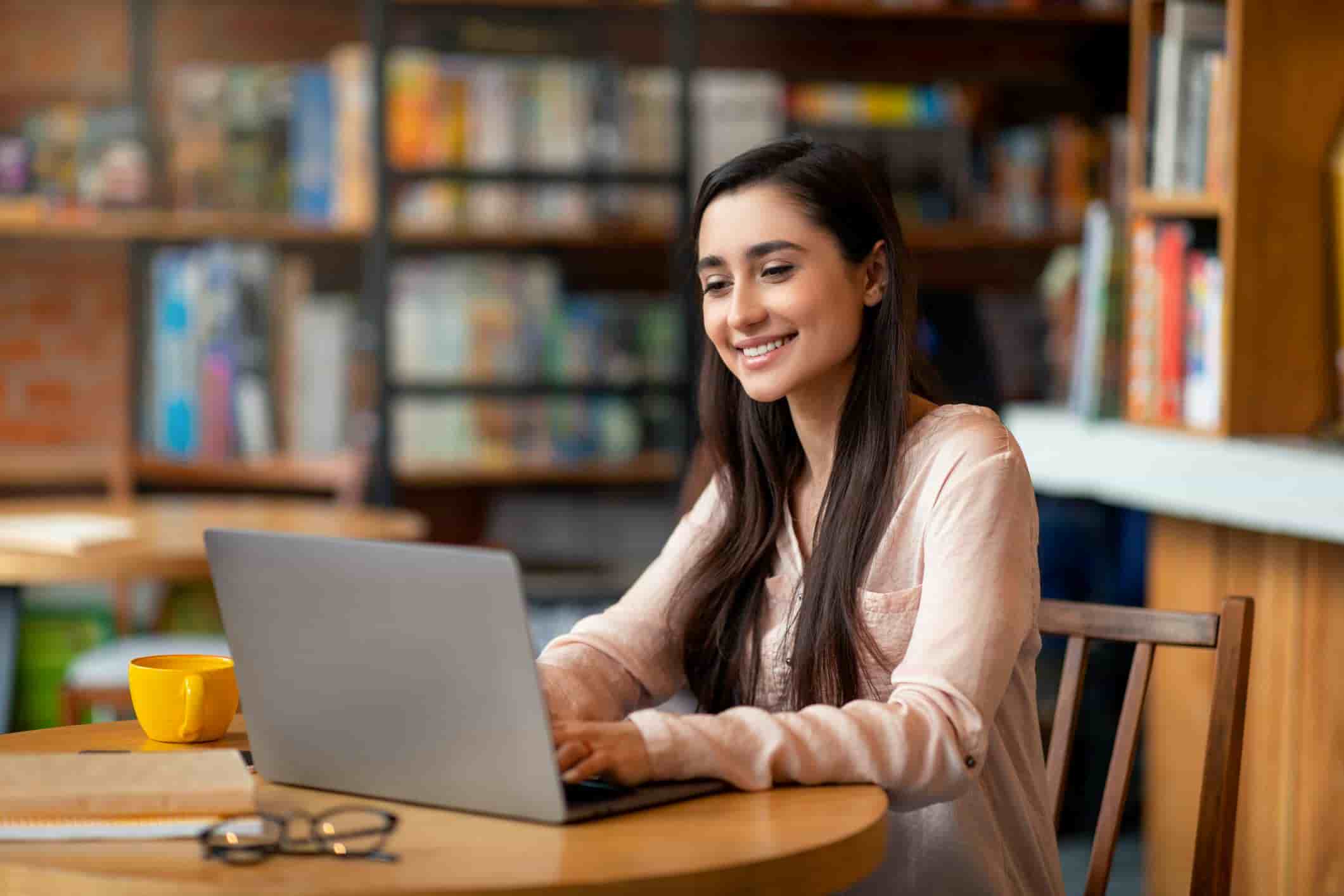 Smiling young woman studying on a laptop in a cozy library or cafe setting, surrounded by bookshelves, with a notebook, glasses, and a yellow coffee cup on the table.