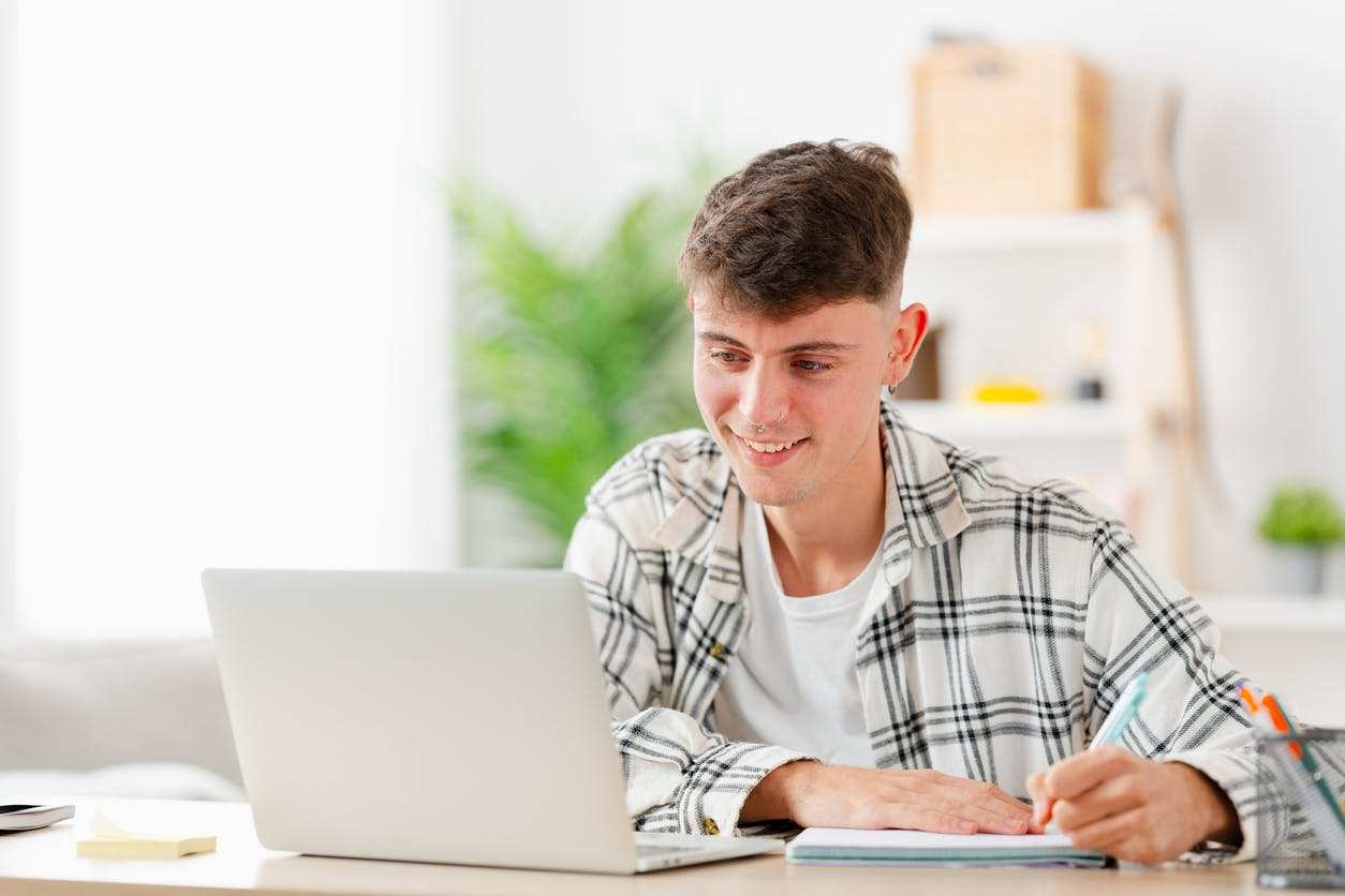 Young student with a nose piercing wearing a plaid shirt, smiling while working on a laptop at home. He is taking notes in a notebook, engaged in studying in a bright, modern workspace with plants in the background