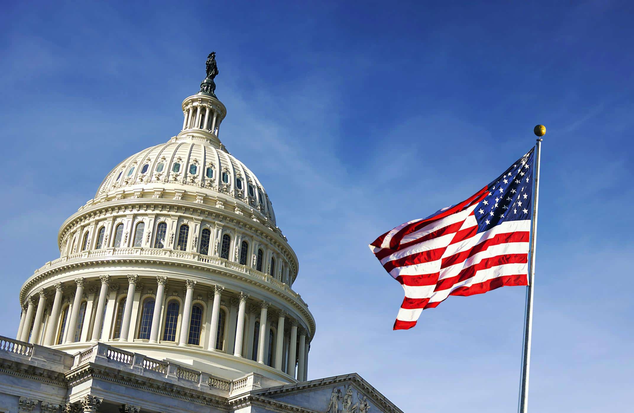 U.S. Capitol Building with American flag waving in the foreground under a clear blue sky.