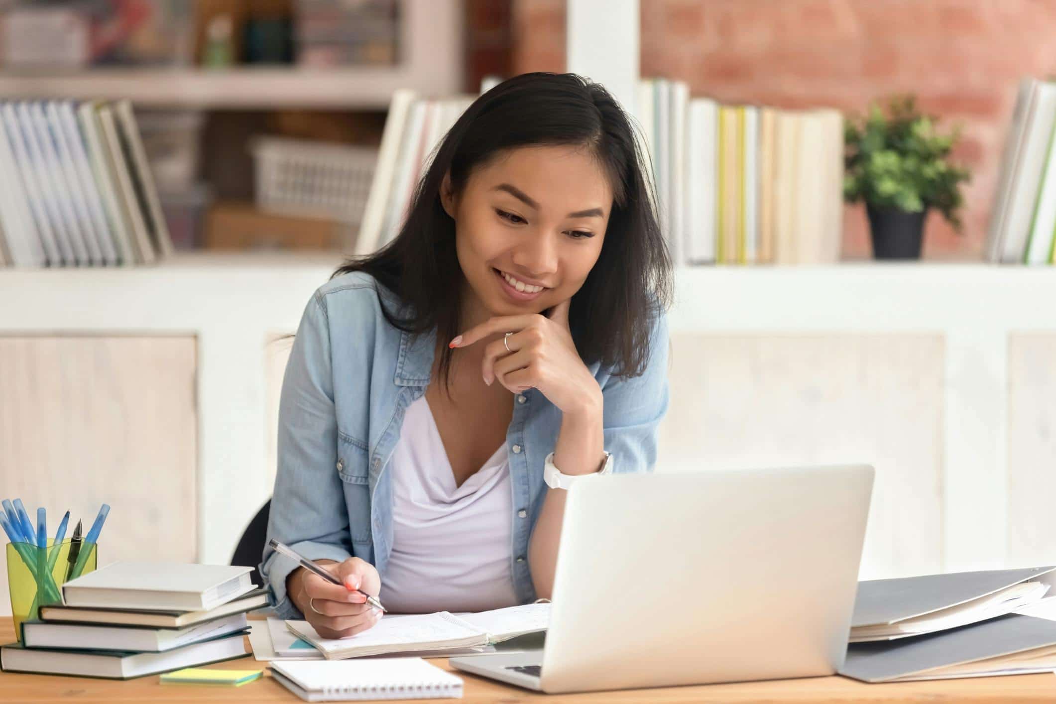 Smiling student studying at home with books, notebooks, and a laptop, engaging in online learning or remote coursework.